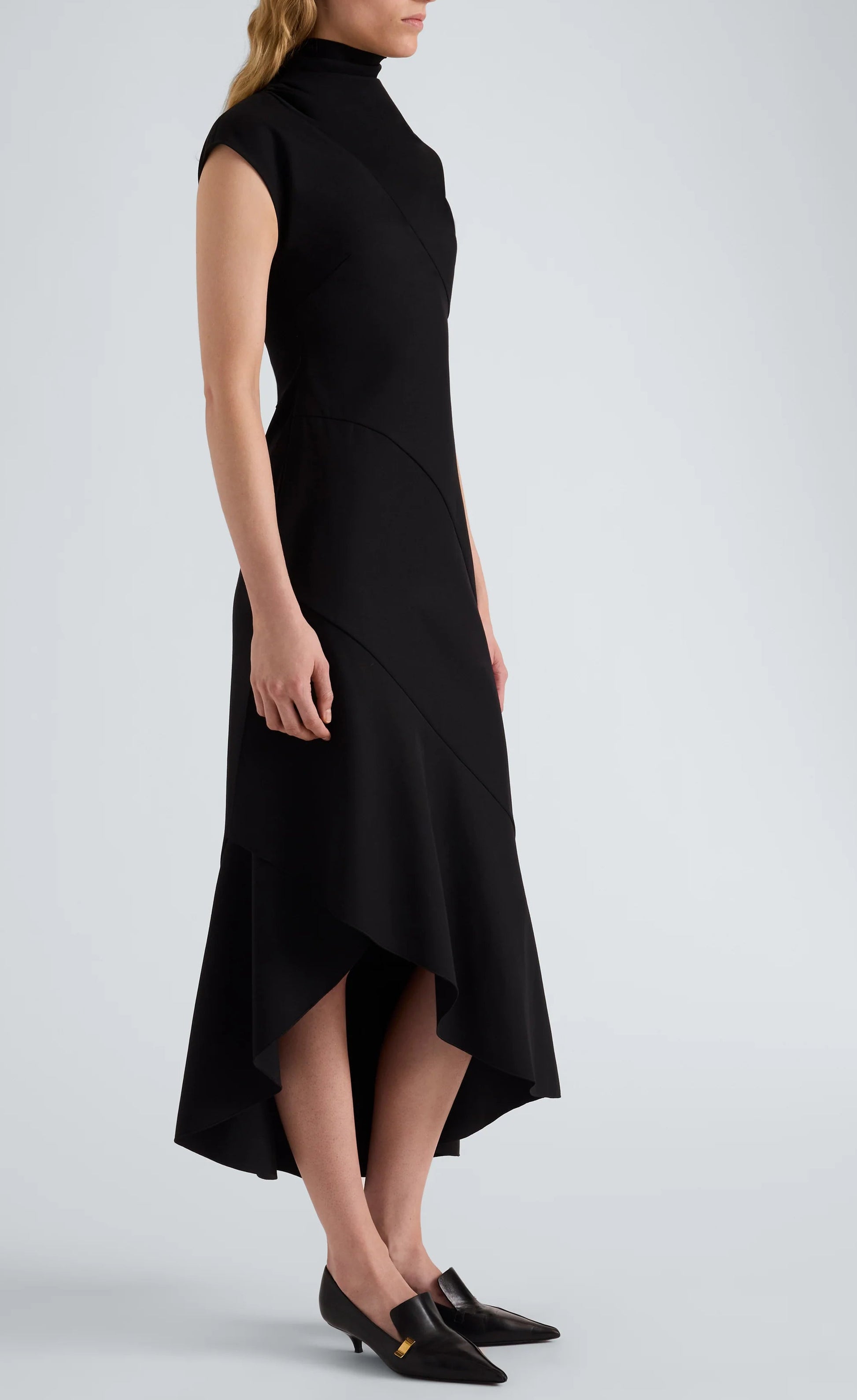 A woman stands against a plain background, modeling the Proenza Schouler Elodine Tropical Wool Dress—a sleeveless, high-neck, asymmetrical black midi crafted from lightweight wool—highlighting its sleek transitional silhouette paired with black pointed-toe shoes.
