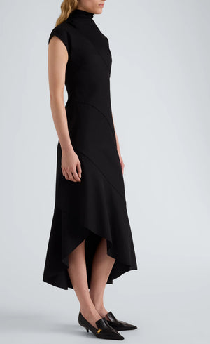 A woman stands against a plain background, modeling the Proenza Schouler Elodine Tropical Wool Dress—a sleeveless, high-neck, asymmetrical black midi crafted from lightweight wool—highlighting its sleek transitional silhouette paired with black pointed-toe shoes.