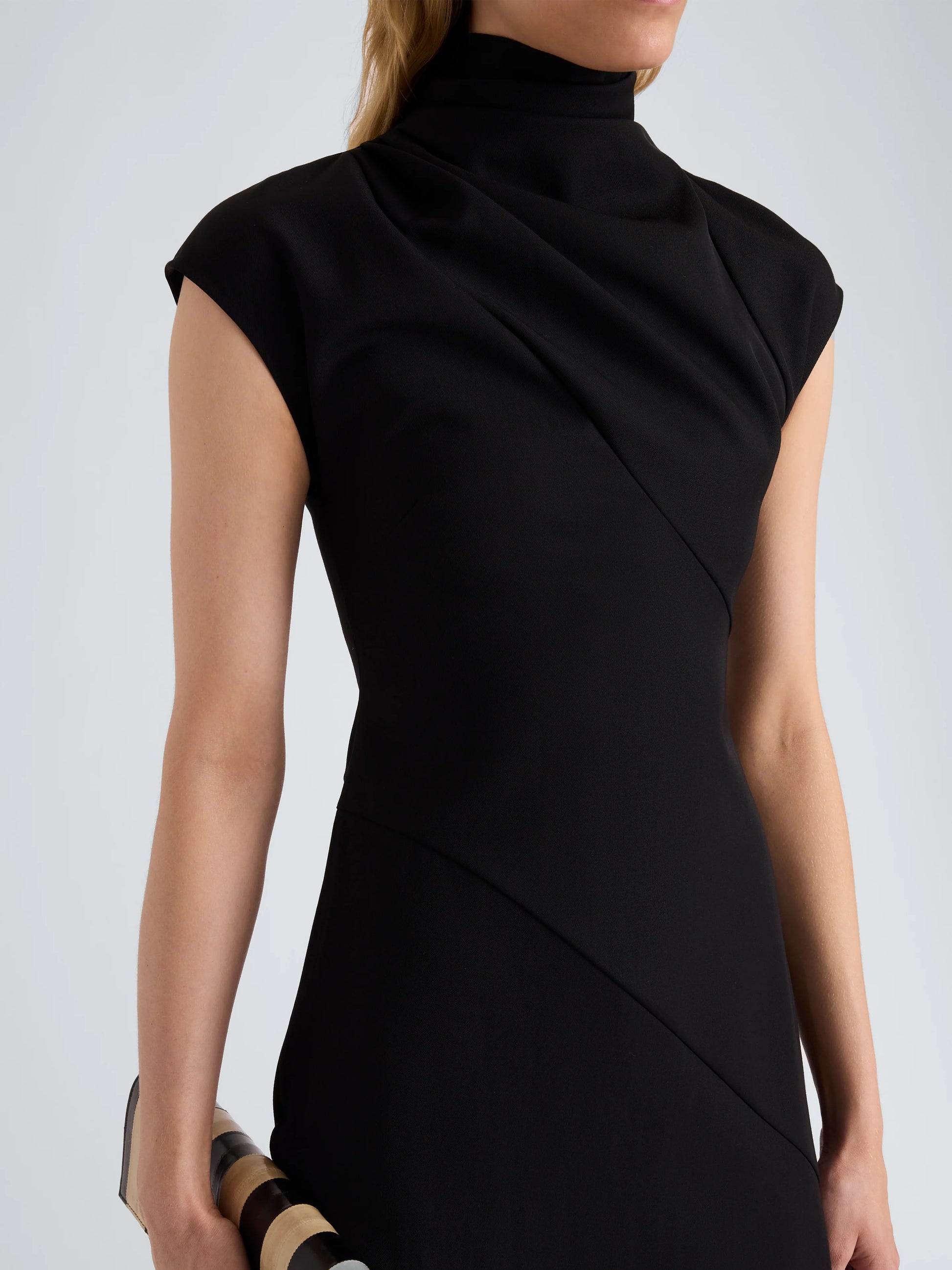 A woman wearing the Proenza Schouler Elodine Tropical Wool Dress—a black, short-sleeve, high-neck style with a transitional silhouette—holds a cylindrical clutch with black and beige stripes.