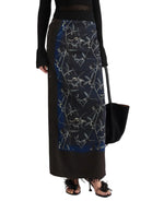 A person wears the Proenza Schouler Anais Skirt—a long, dark skirt adorned with a blue and gray floral pattern inspired by Brazilian night orchids—paired with a black top, heeled sandals, and carrying a black tote bag.