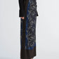 A model wears the Proenza Schouler Anais Skirt in dark floral Printed Stretch Cady with side buttons, styled with a black ribbed sweater featuring flared sleeves and black heeled sandals against a plain background. The look is enriched by the moody elegance of Brazilian night orchids woven throughout the skirt’s print.
