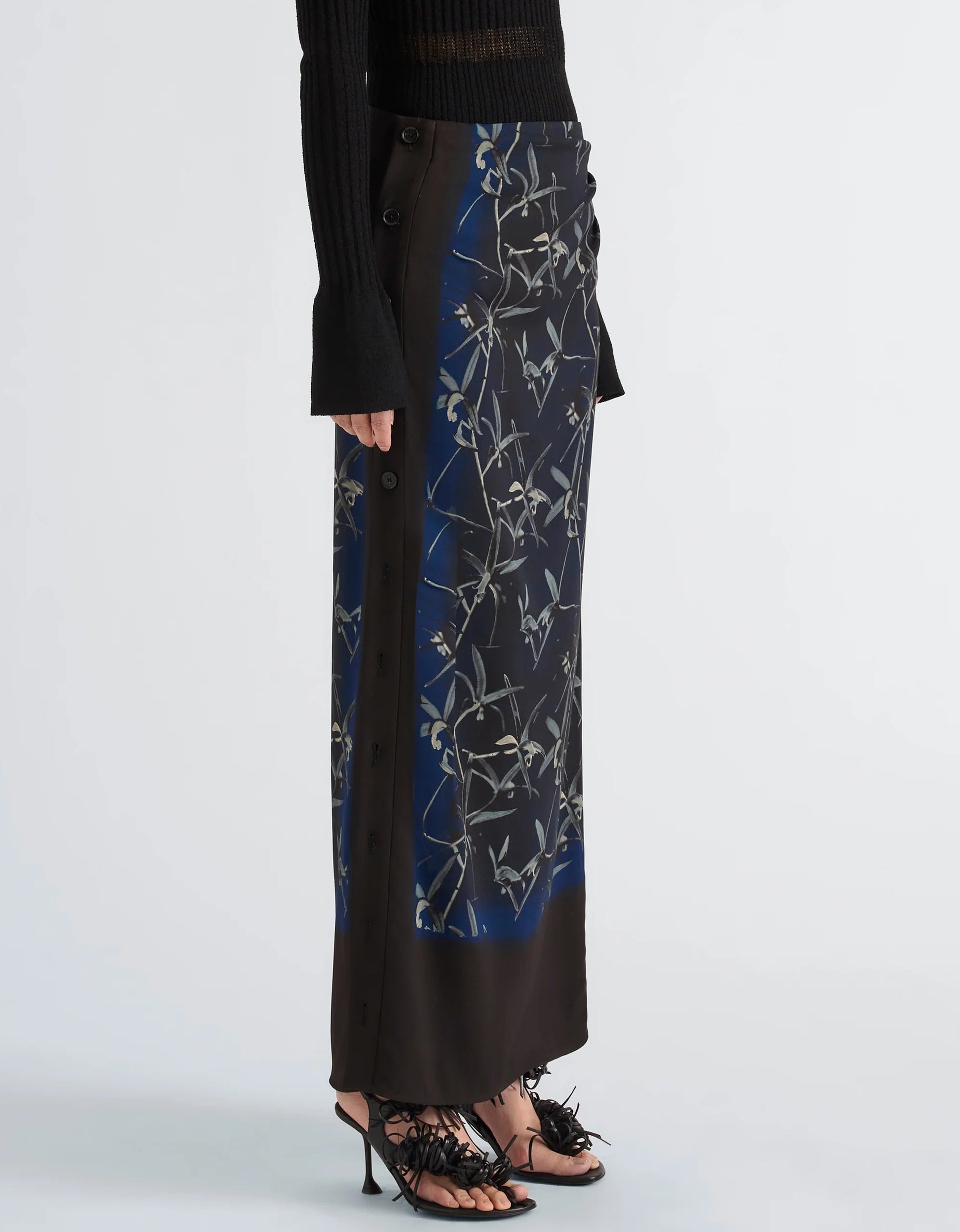 A model wears the Proenza Schouler Anais Skirt in dark floral Printed Stretch Cady with side buttons, styled with a black ribbed sweater featuring flared sleeves and black heeled sandals against a plain background. The look is enriched by the moody elegance of Brazilian night orchids woven throughout the skirt’s print.