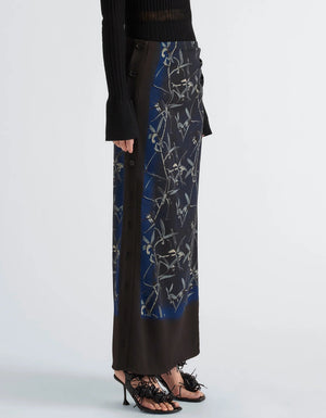 A model wears the Proenza Schouler Anais Skirt in dark floral Printed Stretch Cady with side buttons, styled with a black ribbed sweater featuring flared sleeves and black heeled sandals against a plain background. The look is enriched by the moody elegance of Brazilian night orchids woven throughout the skirt’s print.