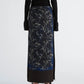 A person is shown from the back against a plain background wearing the Proenza Schouler Anais Skirt in Printed Stretch Cady, which features a striking Brazilian night orchids floral pattern, styled with a black long-sleeve top and black strappy heels.