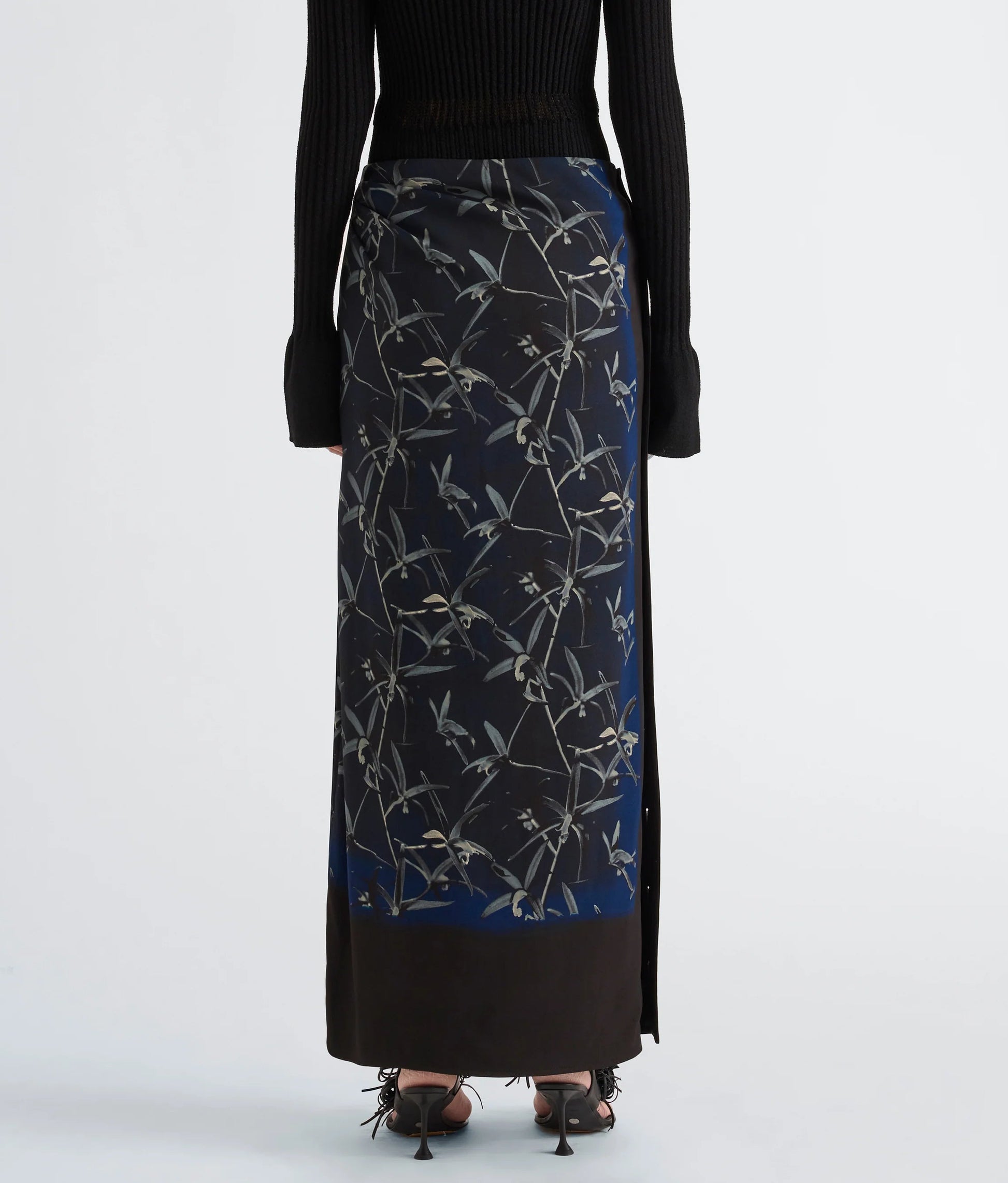A person is shown from the back against a plain background wearing the Proenza Schouler Anais Skirt in Printed Stretch Cady, which features a striking Brazilian night orchids floral pattern, styled with a black long-sleeve top and black strappy heels.