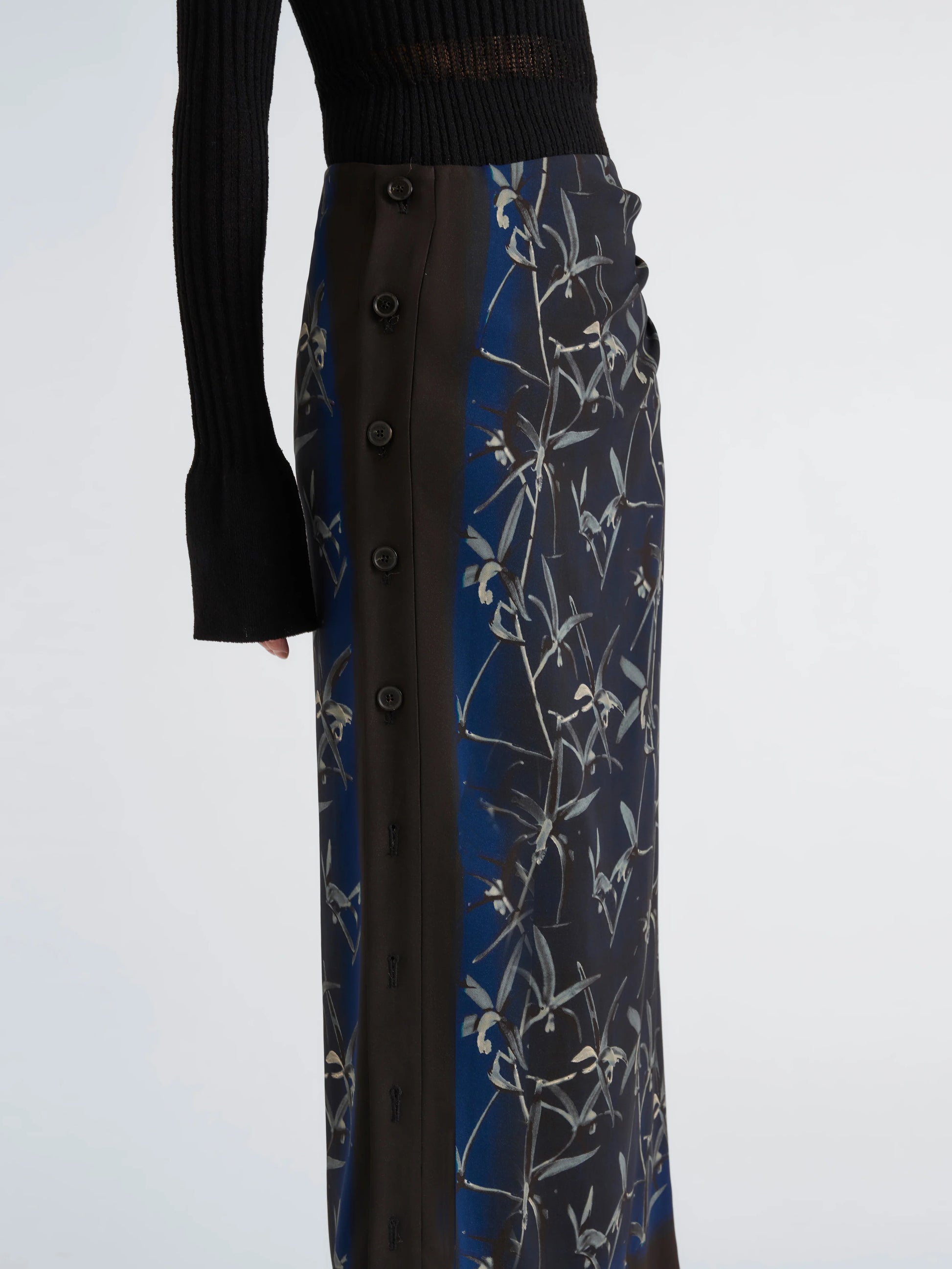 A person wears a black knit top with long sleeves and the Anais Skirt by Proenza Schouler—an ankle-length, slim-fit piece featuring a blue and black floral print reminiscent of Brazilian night orchids.