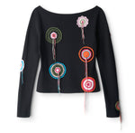 The Rimini Top by Staud is a black slim long-sleeve top with a boat neckline, accented with colorful crocheted circular doily appliqué patches and hanging yarn strands on the front and sleeves.