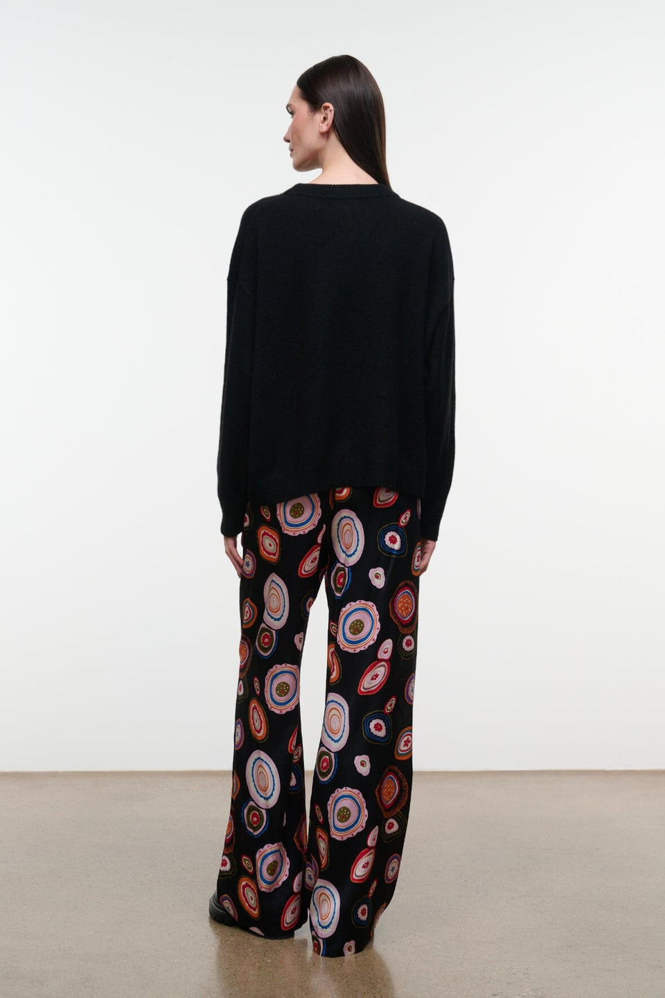 A person stands with their back to the camera, wearing a black sweater paired with Staud’s Stroll Silk Pant—a wide-leg silhouette adorned with a colorful circular pattern that drapes effortlessly against a plain background.