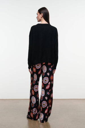 A person stands with their back to the camera, wearing a black sweater paired with Staud’s Stroll Silk Pant—a wide-leg silhouette adorned with a colorful circular pattern that drapes effortlessly against a plain background.