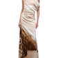 A woman wearing the Staud Phare Silk Maxi Dress, a floor-length silk dress with a one-shoulder neckline and a sepia coastal landscape print, pairs it with black open-toe heels while standing against a white background.