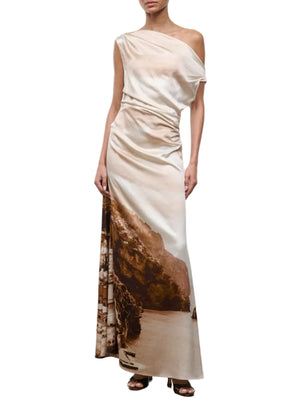A woman wearing the Staud Phare Silk Maxi Dress, a floor-length silk dress with a one-shoulder neckline and a sepia coastal landscape print, pairs it with black open-toe heels while standing against a white background.