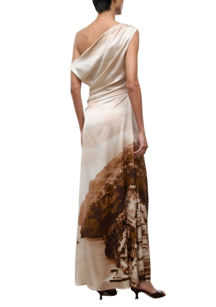 A person wearing the Staud Phare Silk Maxi Dress, featuring a one-shoulder neckline and floor-length silhouette with a printed landscape scene, stands in profile facing away from the camera.