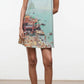 A woman wears the Staud Brio Dress, a pull-on tent silhouette featuring a printed beach scene with people, umbrellas, and water—perfect for warm weather. She completes the look with sandals.