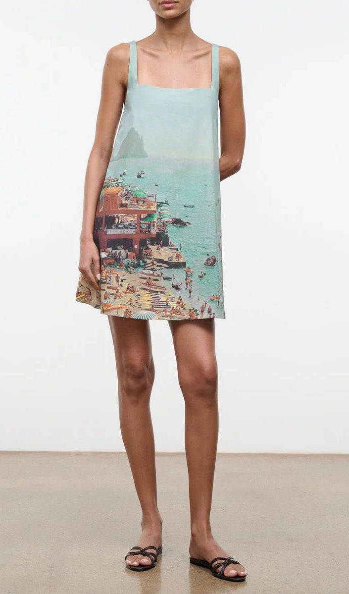 A woman wears the Staud Brio Dress, a pull-on tent silhouette featuring a printed beach scene with people, umbrellas, and water—perfect for warm weather. She completes the look with sandals.