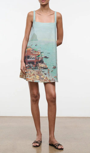 A woman wears the Staud Brio Dress, a pull-on tent silhouette featuring a printed beach scene with people, umbrellas, and water—perfect for warm weather. She completes the look with sandals.