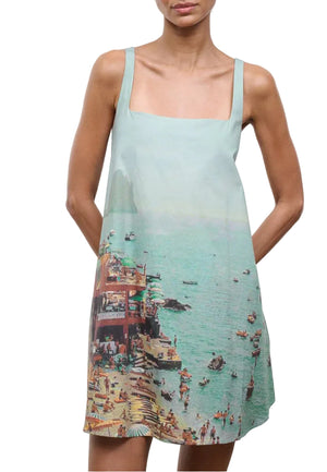 A woman wears the Staud Brio Dress, a sleeveless, square-neck tent silhouette with a printed seaside scene of people, boats, and a pier—an easy pull-on style ideal for warm-weather dressing.