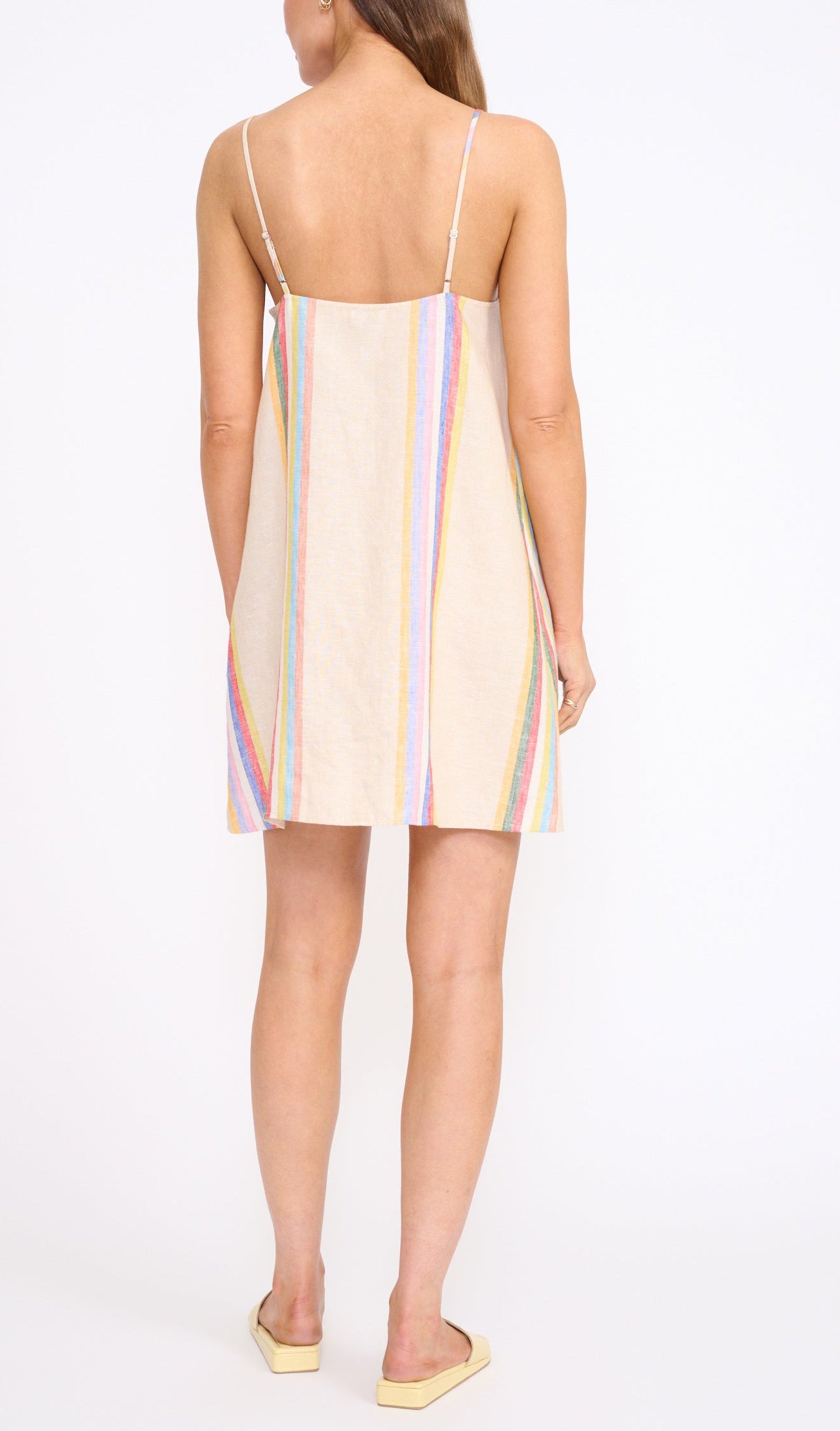 A woman stands facing away in the Staud Laura Linen Mini Dress, featuring summer stripes and spaghetti straps, paired with flat beige shoes against a plain white background.