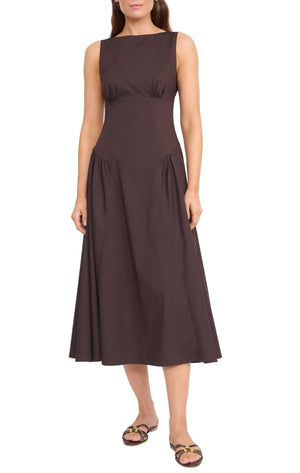 A woman wears the Staud Gwen Dress— a sleeveless, dark brown A-line dress with a fitted bodice, flared skirt, and subtle structured pleats— styled with flat sandals.