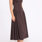 A woman wearing the Staud Gwen Dress—a sleeveless, dark brown A-line midi with a fitted bodice, flared pleated skirt, and flat sandals—stands against a plain white background.