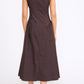 A woman faces away, wearing the Staud Gwen Dress—a sleeveless, dark brown A-line dress with structured pleats—and black sandals against a plain white background.
