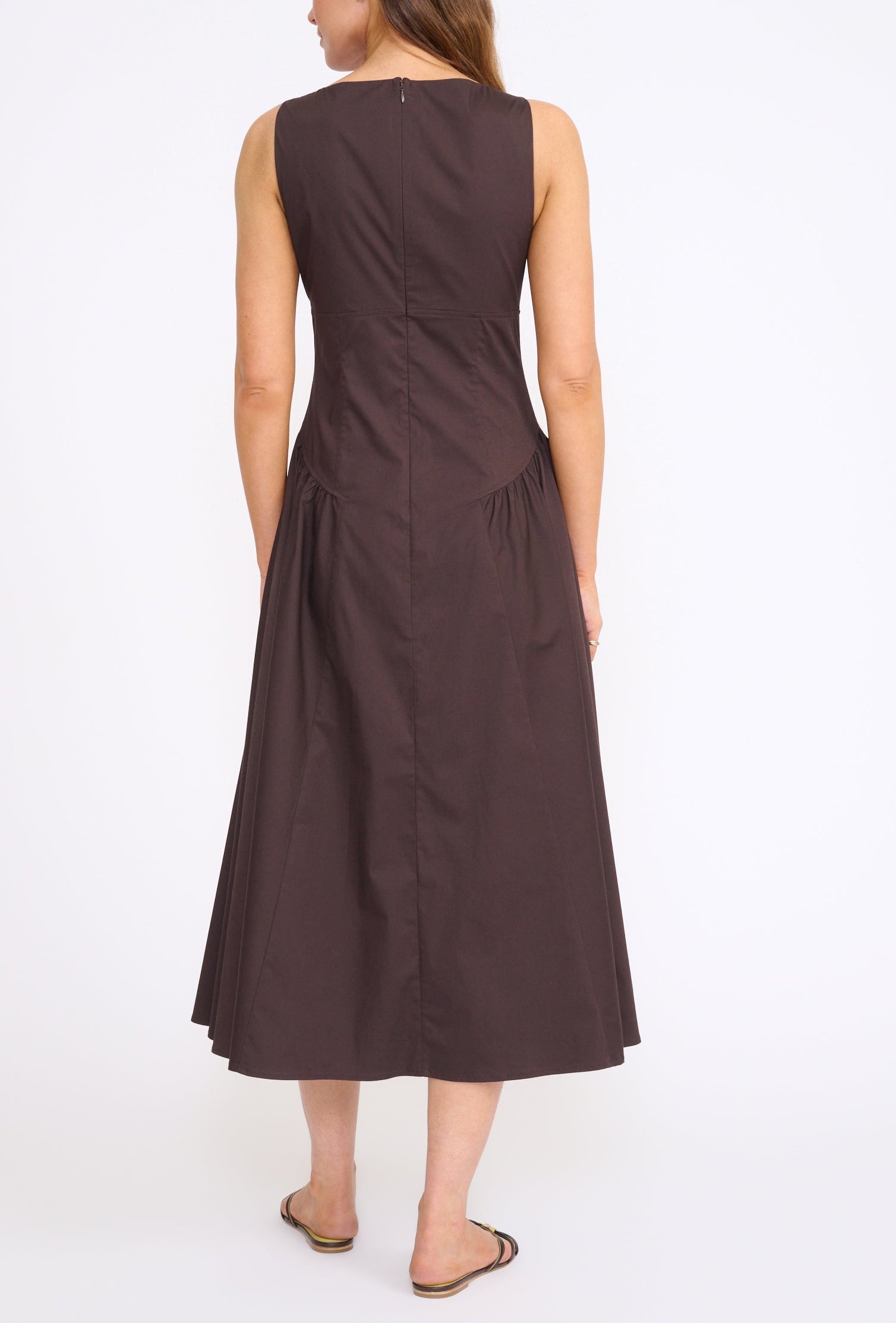 A woman faces away, wearing the Staud Gwen Dress—a sleeveless, dark brown A-line dress with structured pleats—and black sandals against a plain white background.
