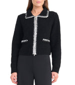 A woman wears the Staud Jeremy Sweater, a black zip-up jacket with white stitched trim on the collar, zipper, and pockets, paired with black pants.