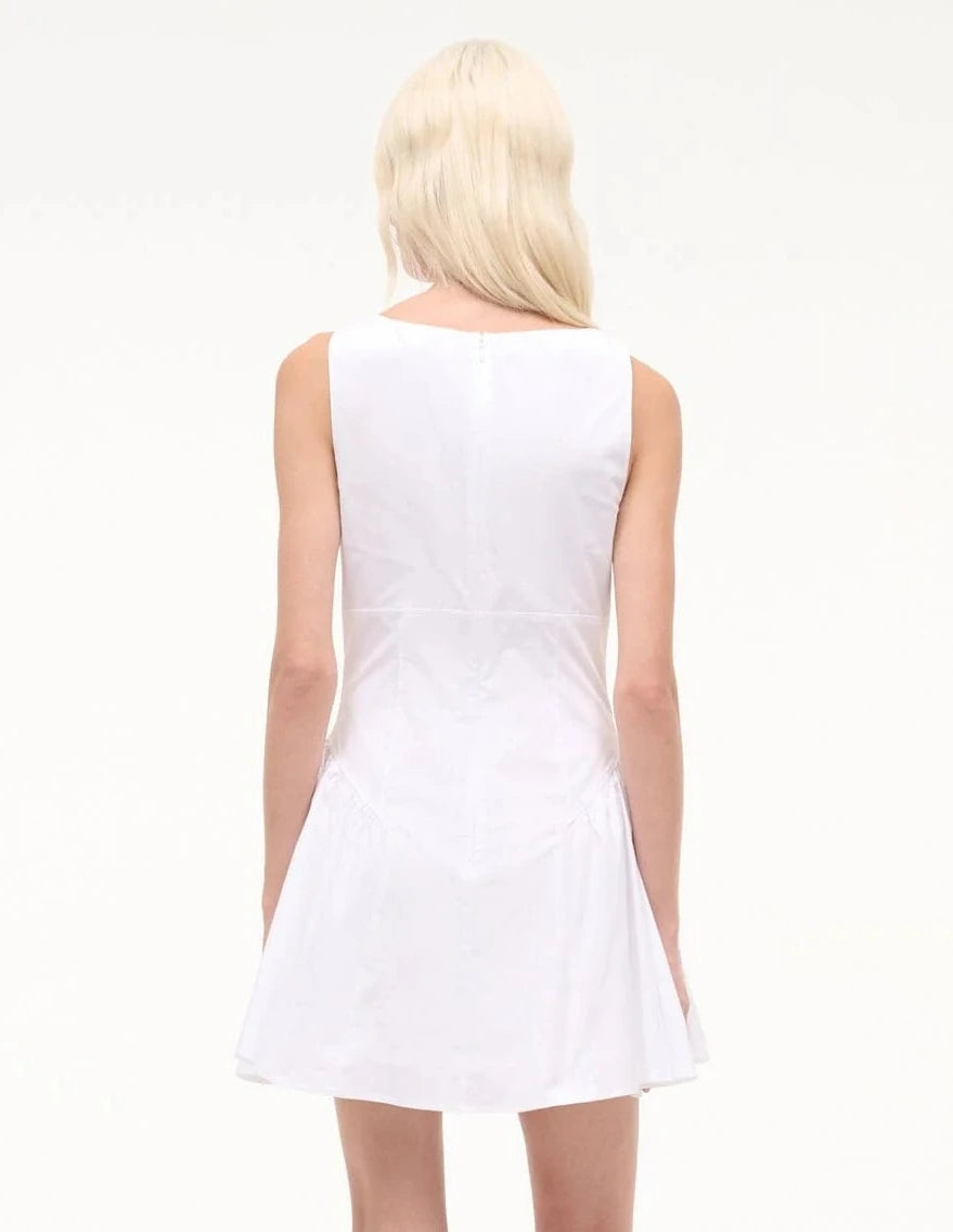 A person with long, light blonde hair is shown from the back, wearing the Staud Gwen Mini Dress—a sleeveless white poplin style with structured pleats—against a plain, light background.