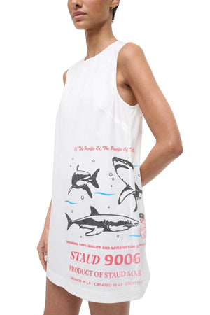 A woman wears the Staud Brea Dress, a sleeveless white shift-style mini dress featuring printed sharks and red text.
