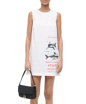 A woman wears the Staud Brea Dress—a sleeveless white mini shift dress with a dolphin graphic and text—while carrying a black textured handbag.