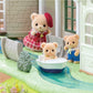 Three Calico Critters bear figures are near a small plastic river and boat in front of the Red Roof Riverside Manor; one bear wears a red dress and hat, another wears blue overalls, and one is sitting in the boat.