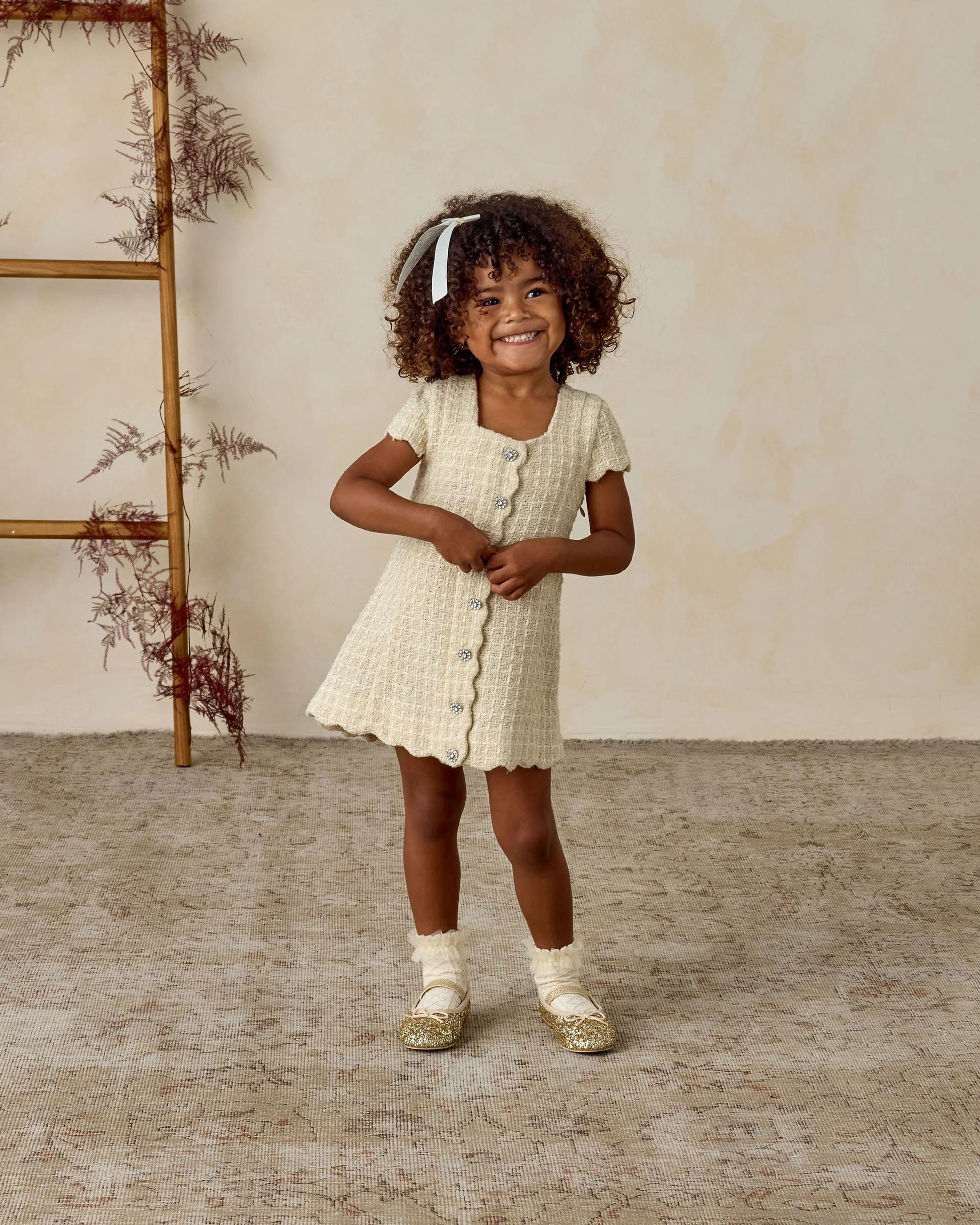 A young girl with curly hair beams on a textured rug, her Noralee Ruffle Socks peeking out beneath a cream cotton-blend dress and gold shoes. A decorative ladder and dried foliage complete the charming scene.