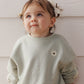 A young child with light brown hair in pigtails and bows wears the Rylee & Cru Baby Girls’ and Girls’ Oversized Sweatshirt, featuring a small flower patch, while standing against a white textured wall.