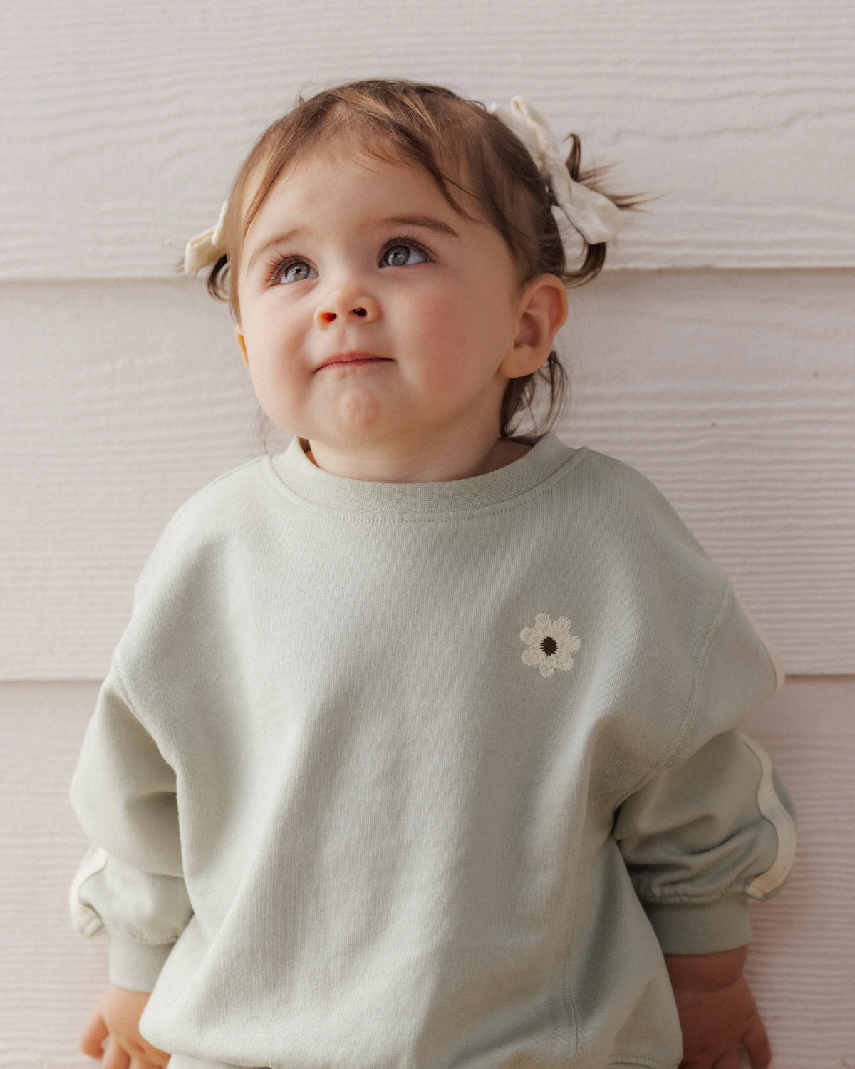 A young child with light brown hair in pigtails and bows wears the Rylee & Cru Baby Girls’ and Girls’ Oversized Sweatshirt, featuring a small flower patch, while standing against a white textured wall.