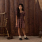A young girl stands indoors on a rug near a wooden chair and pinecones, wearing the Rylee & Cru Girls’ Dillion Dress—a patterned style with a ruffle neckline and smocked bodice—set against a backdrop of brown wooden paneling and burlap banners.
