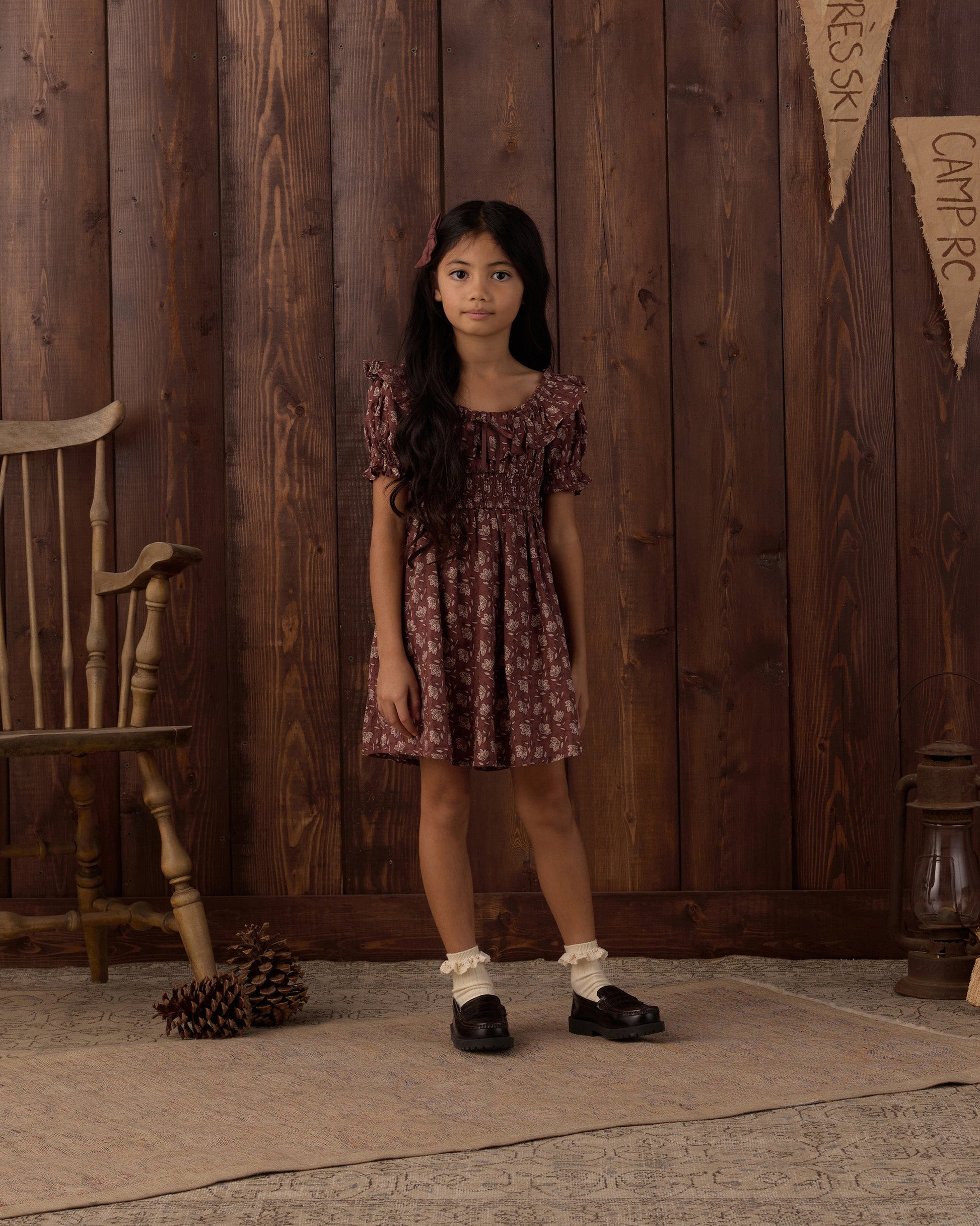 A young girl stands indoors on a rug near a wooden chair and pinecones, wearing the Rylee & Cru Girls’ Dillion Dress—a patterned style with a ruffle neckline and smocked bodice—set against a backdrop of brown wooden paneling and burlap banners.