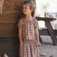 A young girl with curly hair stands outside near a wooden wall and picnic table, wearing the Rylee & Cru Girls’ Ada Blouse—a pink floral smocked top with ruffle sleeves—and looks to the side.