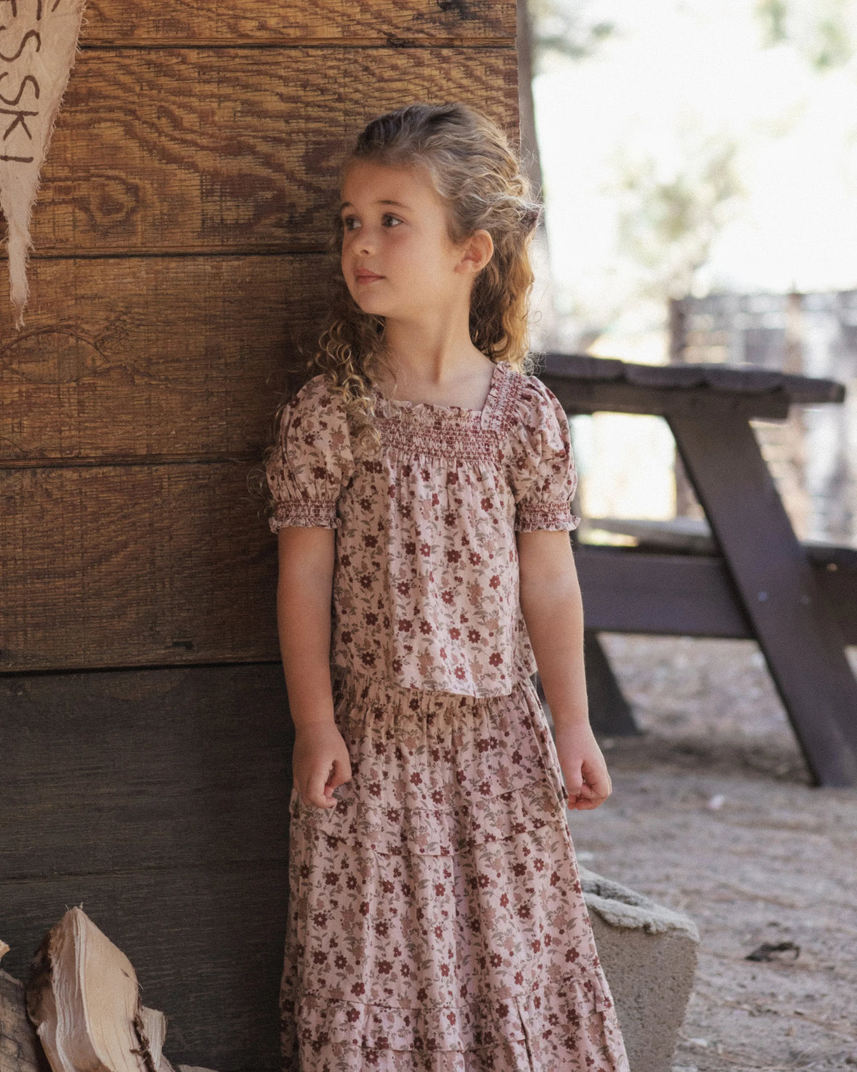 A young girl with curly hair stands outside near a wooden wall and picnic table, wearing the Rylee & Cru Girls’ Ada Blouse—a pink floral smocked top with ruffle sleeves—and looks to the side.