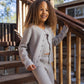A young child with curly hair stands on outdoor wooden stairs, smiling and looking away from the camera while wearing the Rylee & Cru Girls’ Scallop Cardigan in light gray for a cozy layered look.