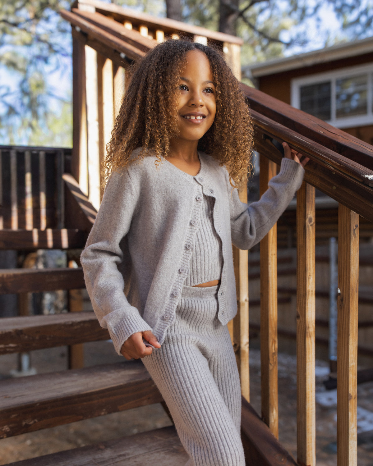 A young child with curly hair stands on outdoor wooden stairs, smiling and looking away from the camera while wearing the Rylee & Cru Girls’ Scallop Cardigan in light gray for a cozy layered look.