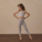 A young girl stands barefoot on a wooden floor, wearing the Rylee & Cru Girls’ Swift Sports Bra with matching leggings in a leopard print classic fit, posing with hands on hips and looking to the side against a beige background—perfect for everyday wear.