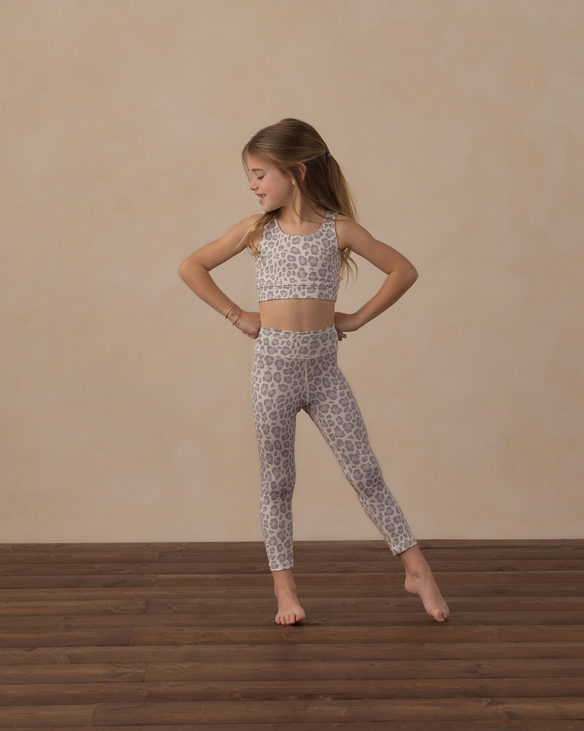A young girl stands barefoot on a wooden floor, wearing the Rylee & Cru Girls’ Swift Sports Bra with matching leggings in a leopard print classic fit, posing with hands on hips and looking to the side against a beige background—perfect for everyday wear.