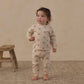 A toddler wearing the Rylee & Cru Baby Girls’ Long Sleeve Tee + Pant Set—featuring a floral-patterned cotton jersey top and matching slouch pants—stands barefoot on a rug beside a rustic wooden stool, facing forward against a neutral background.
