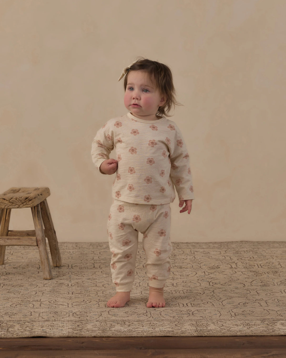 A toddler wearing the Rylee & Cru Baby Girls’ Long Sleeve Tee + Pant Set—featuring a floral-patterned cotton jersey top and matching slouch pants—stands barefoot on a rug beside a rustic wooden stool, facing forward against a neutral background.