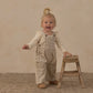 A smiling baby girl wears the Rylee & Cru Baby Girls’ Shoulder Ruffle Bodysuit in an animal-print design, standing next to a small wooden stool on a textured rug with a neutral background.