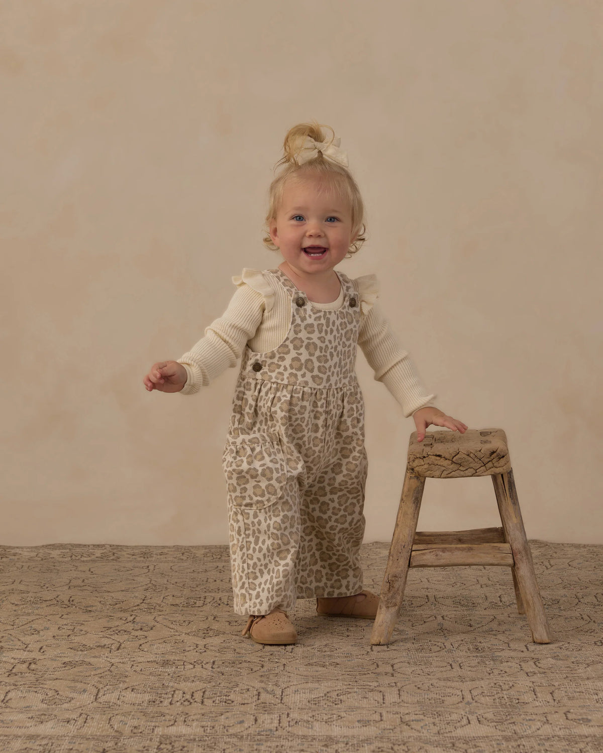 A smiling baby girl wears the Rylee & Cru Baby Girls’ Shoulder Ruffle Bodysuit in an animal-print design, standing next to a small wooden stool on a textured rug with a neutral background.