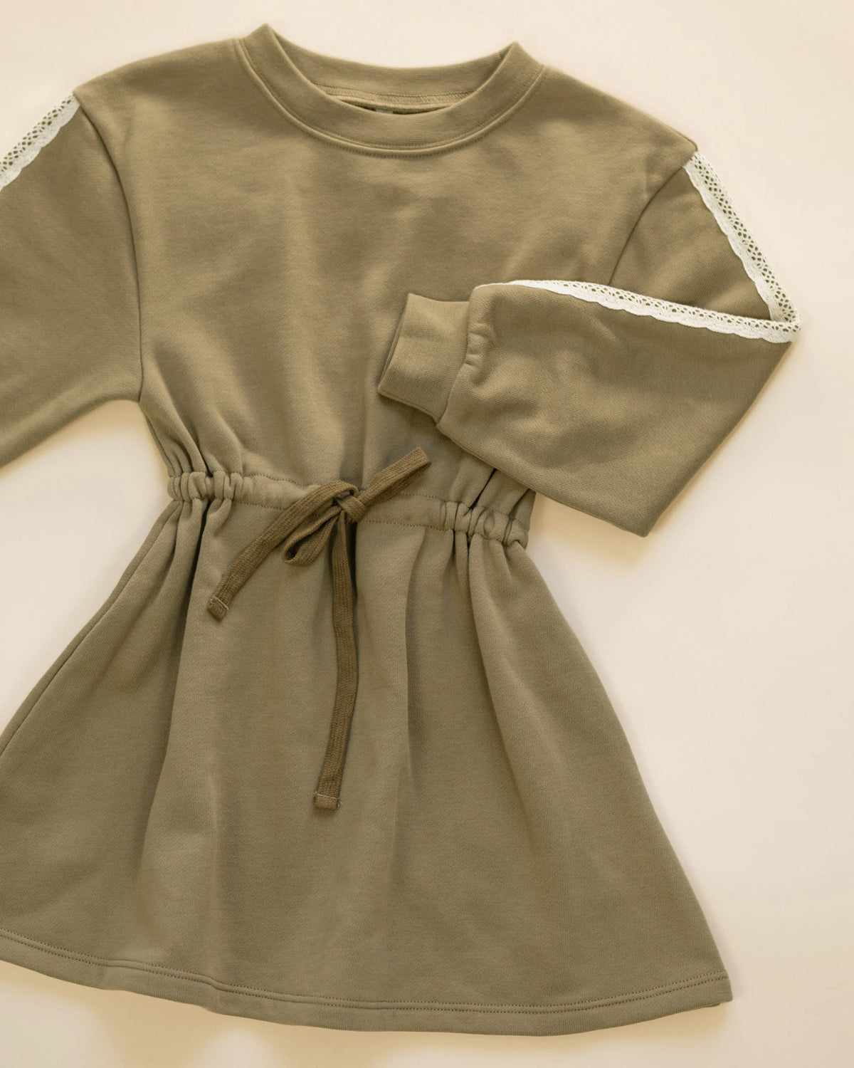 The Rylee & Cru Girls’ Tate Dress in olive green, featuring long sleeves with delicate lace trim, an adjustable drawstring, and a gathered waist, is shown laid flat on a light background.