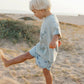 A young blond boy wears Rylee & Cru Baby Boys' & Boys' Swim Trunk as he kicks sand barefoot on a sunny beach with ocean and shrubs in the background. These swim trunks offer UPF 50+ protection for safe, sun-filled play.