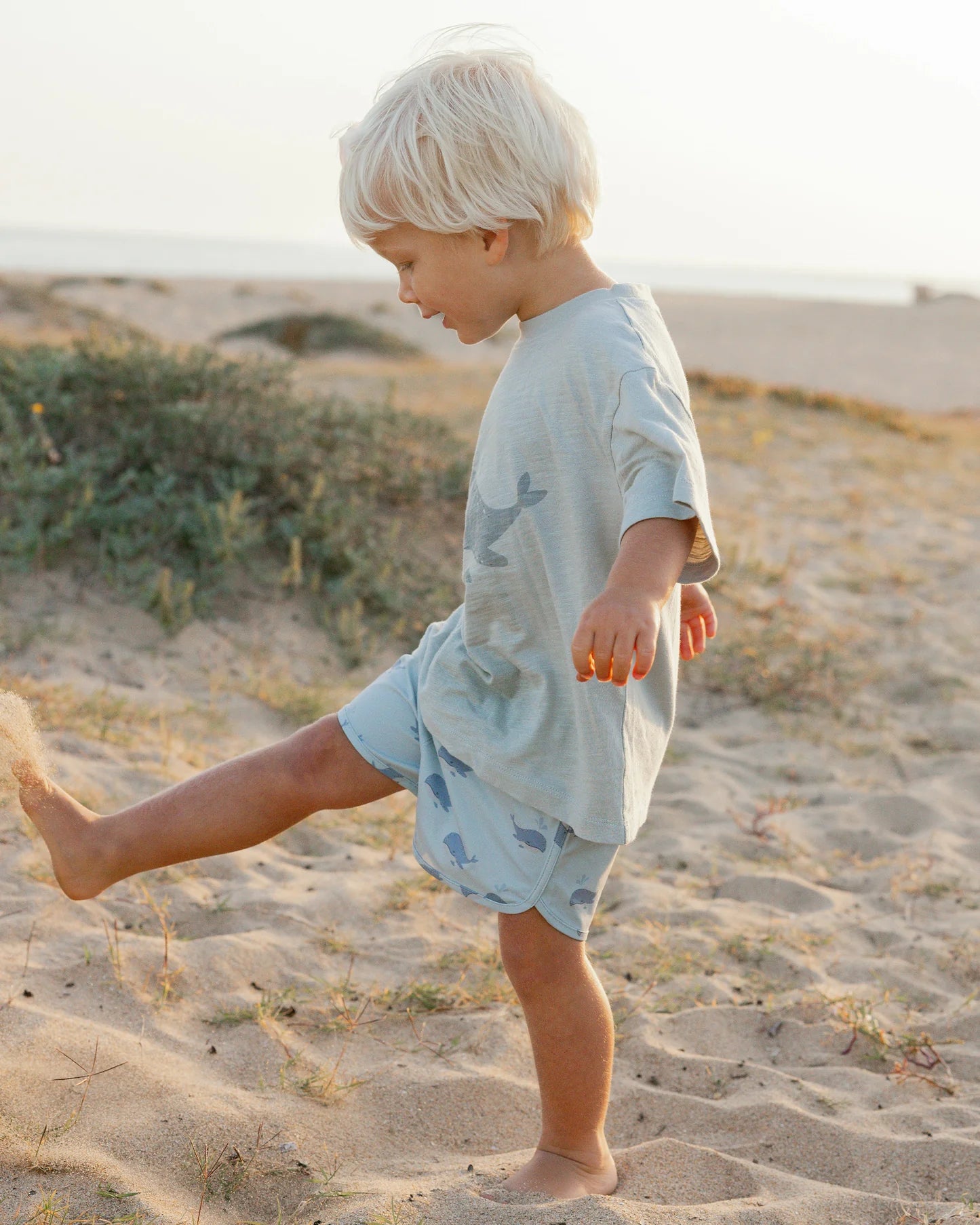 A young blond boy wears Rylee & Cru Baby Boys' & Boys' Swim Trunk as he kicks sand barefoot on a sunny beach with ocean and shrubs in the background. These swim trunks offer UPF 50+ protection for safe, sun-filled play.