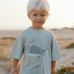 A young child with blond hair stands on sandy, grassy terrain wearing the Rylee & Cru Baby Boys' & Boys' Relaxed Tee, a soft cotton jersey shirt featuring a whale graphic.