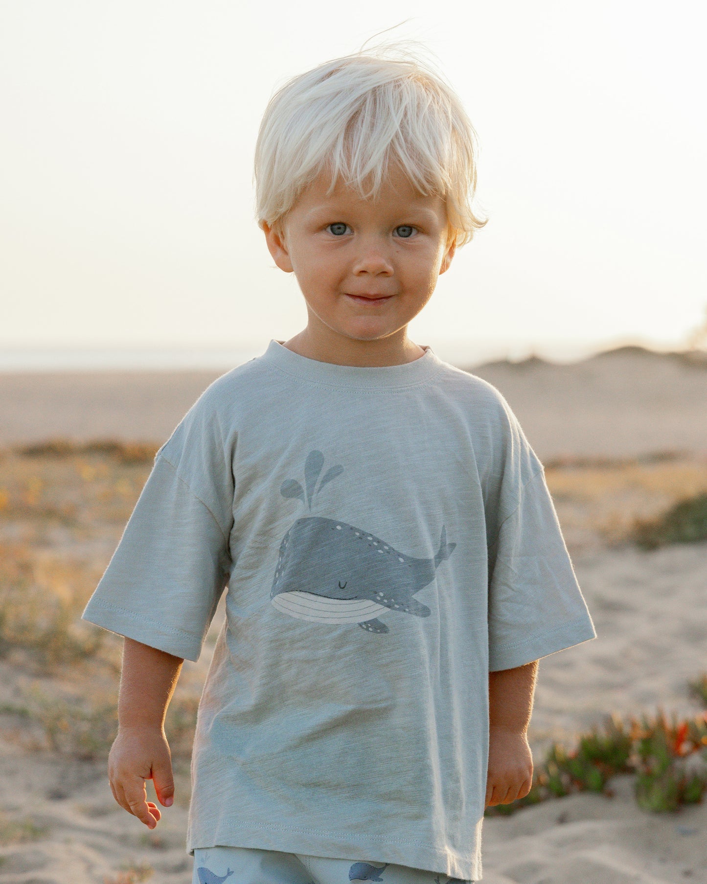A young child with blond hair stands on sandy, grassy terrain wearing the Rylee & Cru Baby Boys' & Boys' Relaxed Tee, a soft cotton jersey shirt featuring a whale graphic.
